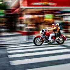 man and woman on a red motorcycle, as they speed through an intersection on a shop filled street.