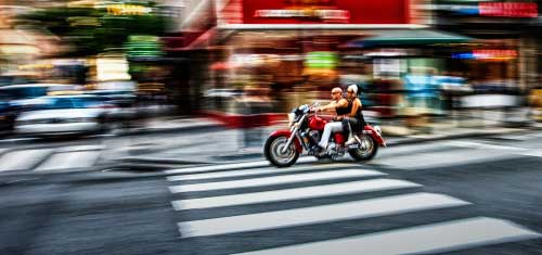 man and woman on a red motorcycle, as they speed through an intersection on a shop filled street.