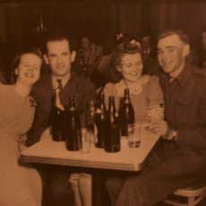 Two couples in the 1940's, wearing suits and dresses, seated at a table with beer bottles on it.