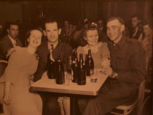 Two couples in the 1940's, wearing suits and dresses, seated at a table with beer bottles on it.