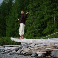 A man balances on a suspended log with bare feet, arms extended, palms raised to the horizon. The BC beach is the background.