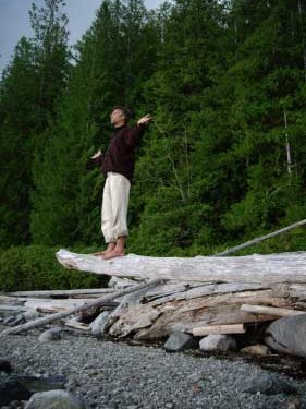 A man balances on a suspended log with bare feet, arms extended, palms raised to the horizon. The BC beach is the background.