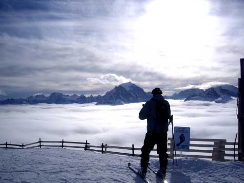 A lone skier is silhouetted overlooking a vast distance to the far mountain range, while standing above the clouds