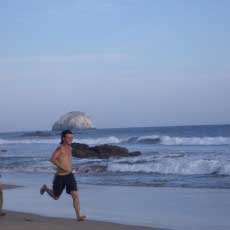 A very fit and happy young couple are sprinting along the seashore with waves crashing in the background.