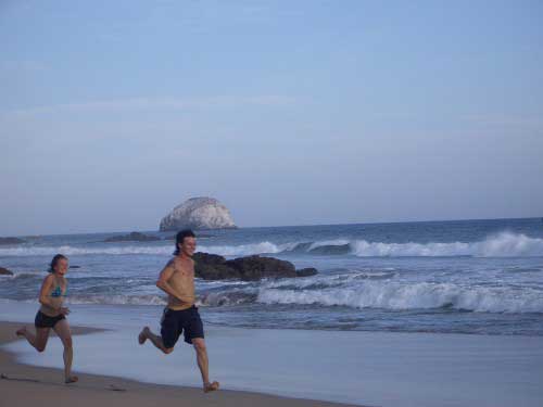 A very fit and happy young couple are sprinting along the seashore with waves crashing in the background.