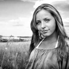 A beautiful woman with minimalist makeup, looks candidly at the camera while standing amid shoulder height grasses with more fields in the distance.