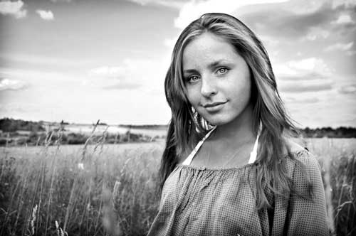 A beautiful woman with minimalist makeup, looks candidly at the camera while standing amid shoulder height grasses with more fields in the distance.