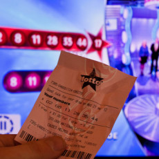 A lottery ticket in a man's hand, comparing to the numbers on the TV screen in the background.