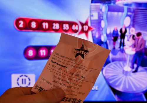 A lottery ticket in a man's hand, comparing to the numbers on the TV screen in the background.