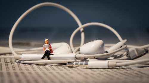 A tiny figure sits on earbuds reading a book, as a depiction of audio books.