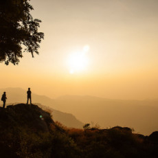 Two silhouetted people look to the distant mountains at sunset. The sky is many shades of pale orange; a monochrome.