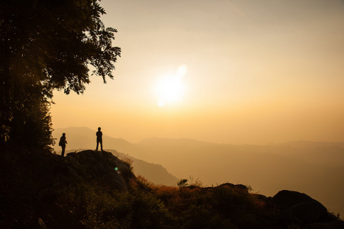 Two silhouetted people look to the distant mountains at sunset. The sky is many shades of pale orange; a monochrome.