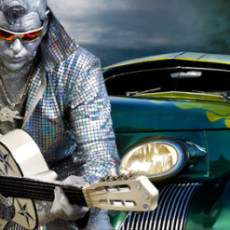 A man imitates Elvis, dressed all in sparkly silver with silver face paint leans on an old car while holding a guitar.