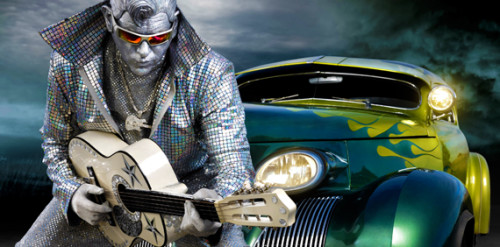 A man imitates Elvis, dressed all in sparkly silver with silver face paint leans on an old car while holding a guitar.