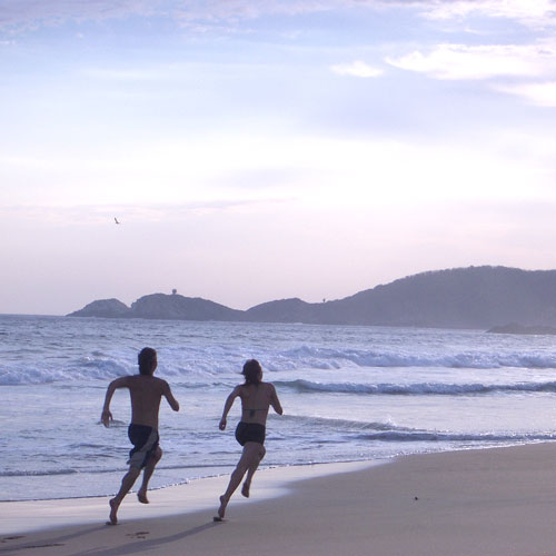 A healthy woman and man sprint along the beach toward the distant mountains. No gear workouts suit a minimalist lifestyle.